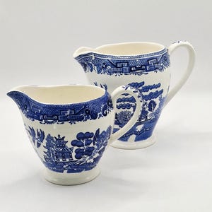 May include: Two white ceramic cream pitchers with blue Willow pattern designs. The pitchers have a classic shape with spouts and handles. The blue and white pattern features pagodas, trees, and bridges, creating a traditional aesthetic.