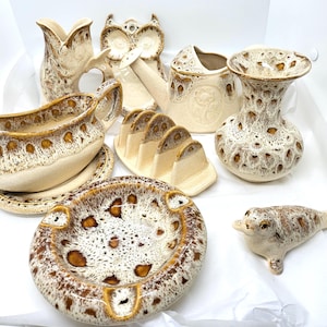 May include: A collection of vintage ceramic pieces in cream and brown tones. The assortment includes a vase, owl-shaped wall decor, a watering can, a gravy boat with a saucer, a toast rack, an ashtray, a decorative vase, and a seal figurine.
