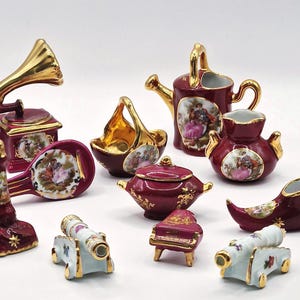 May include: A collection of miniature porcelain figurines in burgundy, gold, and light blue. The set includes a gramophone, watering can, shoe, piano, cannons, and other decorative pieces, each with a painted scene.