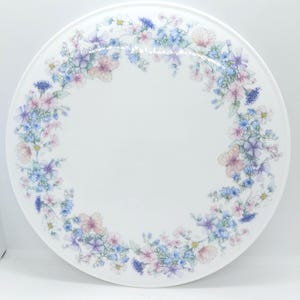 以下が含まれることがあります： 白い丸い装飾皿、花の模様。 皿は、青、ピンク、紫の花を含むパステルカラーの花のボーダーが特徴です。 花は、皿の端の周りに円形に配置されています。