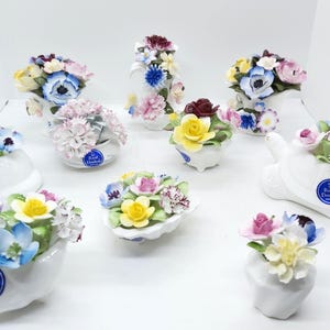 May include: A collection of white porcelain flower figurines, each adorned with colorful, detailed floral arrangements. The figurines are in various shapes, including vases, a cat, a turtle, and shell-shaped bowls.