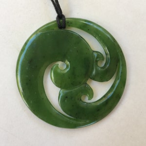 May include: A green jade pendant with a traditional Maori design, known as a koru, which symbolizes new beginnings and growth. The pendant is round and has a black cord attached to it.