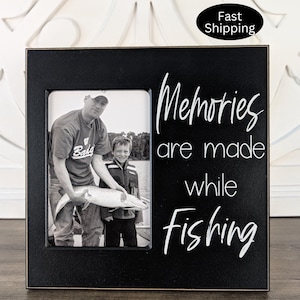 May include: Black wooden picture frame with a white inscription that reads "Memories are made while fishing". A black and white photo of two people fishing is in the frame.