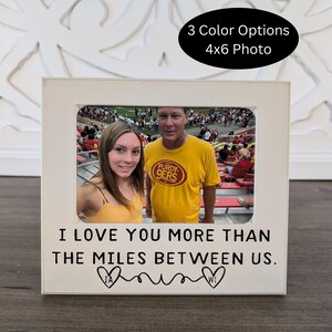 May include: A white photo frame with a picture of two people. The frame has the text "I LOVE YOU MORE THAN THE MILES BETWEEN US." and the words "IA" and "WI" inside hearts. The frame is designed for a 4x6 photo and has "3 Color Options" text.