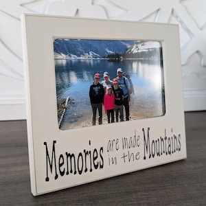 May include: White wooden picture frame with a black inscription that reads "Memories are made in the Mountains". The frame holds a photo of a family of five standing in front of a lake and mountains.