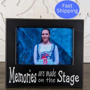 May include: Black wooden picture frame with a white inscription that reads "Memories are made on the Stage". The frame contains a photo of a young woman in a white and green dress standing on a stage.