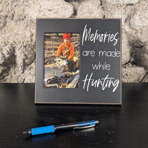 May include: Black framed photo with a picture of a person and a deer. The frame has the text "Memories are made while Hunting" in white script. A blue and black pen rests in front of the frame.