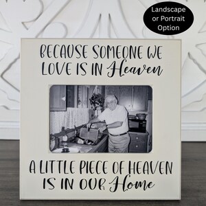 May include: A square white picture frame with black text that reads, "BECAUSE SOMEONE WE LOVE IS IN HEAVEN A LITTLE PIECE OF HEAVEN IS IN OUR HOME." The frame contains a black and white photograph of an older person standing in a kitchen.