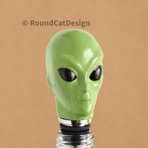 May include: Green alien head bottle stopper with black eyes. The stopper is made of ceramic and has a metal base. It is inserted into a clear glass bottle.