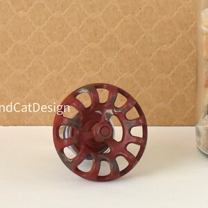 May include: A red metal knob with a circular design and a silver center. The knob has a distressed look with some rust spots.