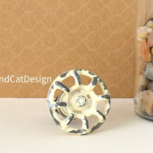 May include: A white and black vintage-style cabinet knob with a distressed finish. The knob has a round shape and a circular design with spokes.