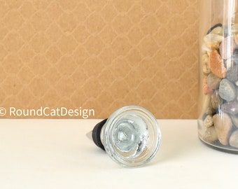 May include: A clear glass bottle stopper with a black cork and a silver ring. The stopper is sitting on a white surface.