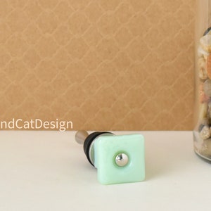 May include: A light green square bottle stopper with a silver knob. The stopper is made of glass and has a smooth, polished finish.