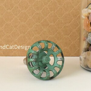 May include: Green metal cabinet knob with a circular design and a decorative, openwork pattern. The knob has a silver stem.