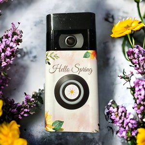 Puede incluir: Una cámara de video negra con un diseño floral que dice "Hello Spring". La cámara está rodeada de flores amarillas y moradas. La cámara tiene una lente y un diseño floral decorativo.