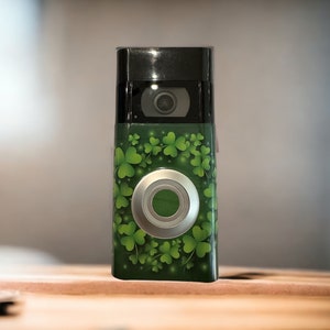 May include: A black and green video doorbell with a shamrock design. The doorbell has a silver button and a camera lens. The green background is covered in light green shamrocks. The doorbell is on a wooden surface.