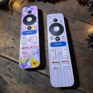 Op de afbeelding: Twee afstandsbedieningen voor de televisie met verschillende ontwerpen. De linker afstandsbediening heeft een kleurrijk bloemenpatroon met paarse, roze en gele bloemen. De rechter afstandsbediening heeft een wit houtnerfpatroon. Beide hebben knoppen voor Free TV, Netflix en YouTube.