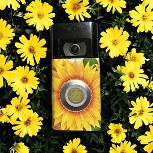 May include: A black security camera with a sunflower design on the front, surrounded by bright yellow flowers with green foliage. The camera has a silver-colored ring around the lens.