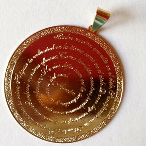 May include: Gold-toned pendant with a circular design featuring a spiral of text in a foreign language. The pendant has a loop at the top for attaching to a chain.
