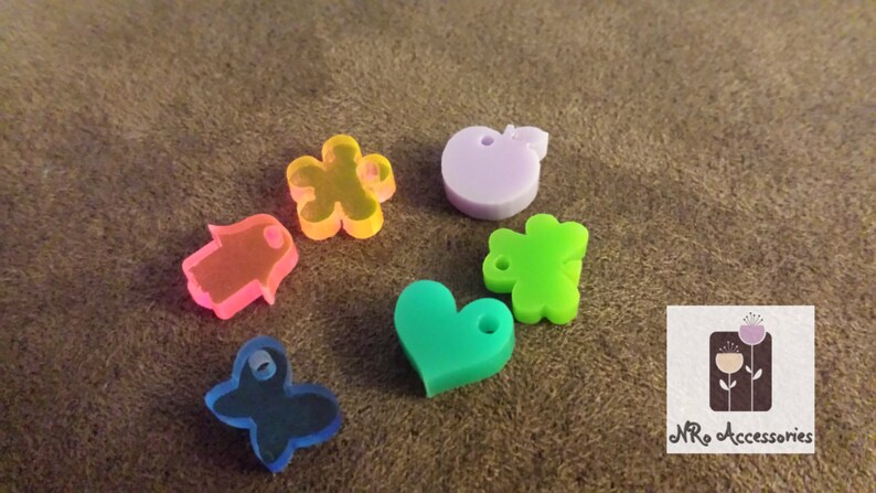 May include: Assortment of colorful plastic charms in various shapes, including a heart, a star, a hand, a bear, an apple, and a butterfly. The charms have holes for attaching to jewelry or other crafts.