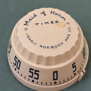 May include: A vintage white kitchen timer with blue numerals and text. The timer reads "Maid of Honor TIMER SEARS, ROEBUCK AND CO." The timer has a ridged edge and a dial that goes up to 55 minutes.