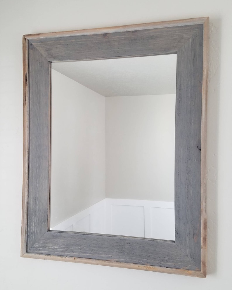 May include: A rectangular mirror with a grey wooden frame. The frame has a distressed, weathered appearance with visible wood grain. The mirror reflects a white wall and a corner of a room.