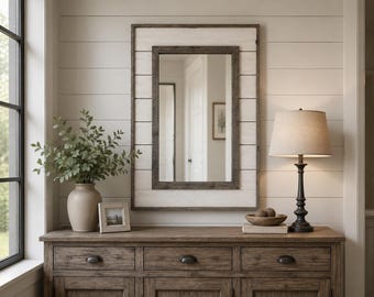 May include: A rectangular wall mirror featuring a dark brown wooden frame set against a white horizontal plank backing. The mirror is positioned above a wooden console table with three drawers, a beige lamp, a ceramic vase with greenery, and a small frame.