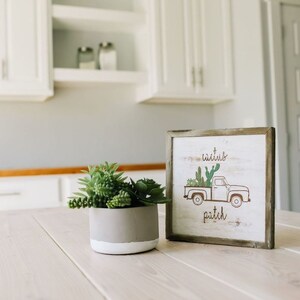 May include: A wooden sign with a white background and brown lettering. The sign features a cartoon image of a white pickup truck with a bed full of cacti. The text on the sign reads "cactus patch". The sign is sitting on a white wooden table with a potted succulent plant in front of it.