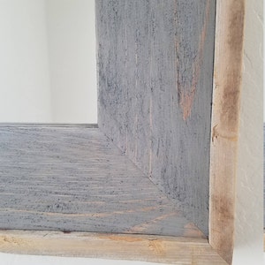 May include: Close-up of a rustic wooden frame with a grey painted finish. The frame has a natural wood edge and a corner joint. The wood grain is visible, adding texture. The frame is likely for a mirror or artwork.