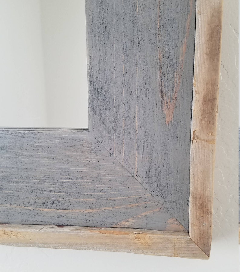 May include: Close-up of a rustic wooden frame with a grey painted finish. The frame has a natural wood edge and a corner joint. The wood grain is visible, adding texture. The frame is likely for a mirror or artwork.