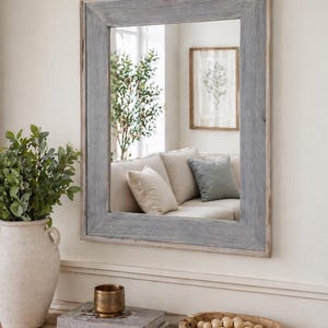 May include: A rectangular mirror with a grey wooden frame hangs on a white wall. The mirror reflects a living room with a sofa and artwork. A vase with greenery, books, and a decorative bowl sit on a wooden table below the mirror.