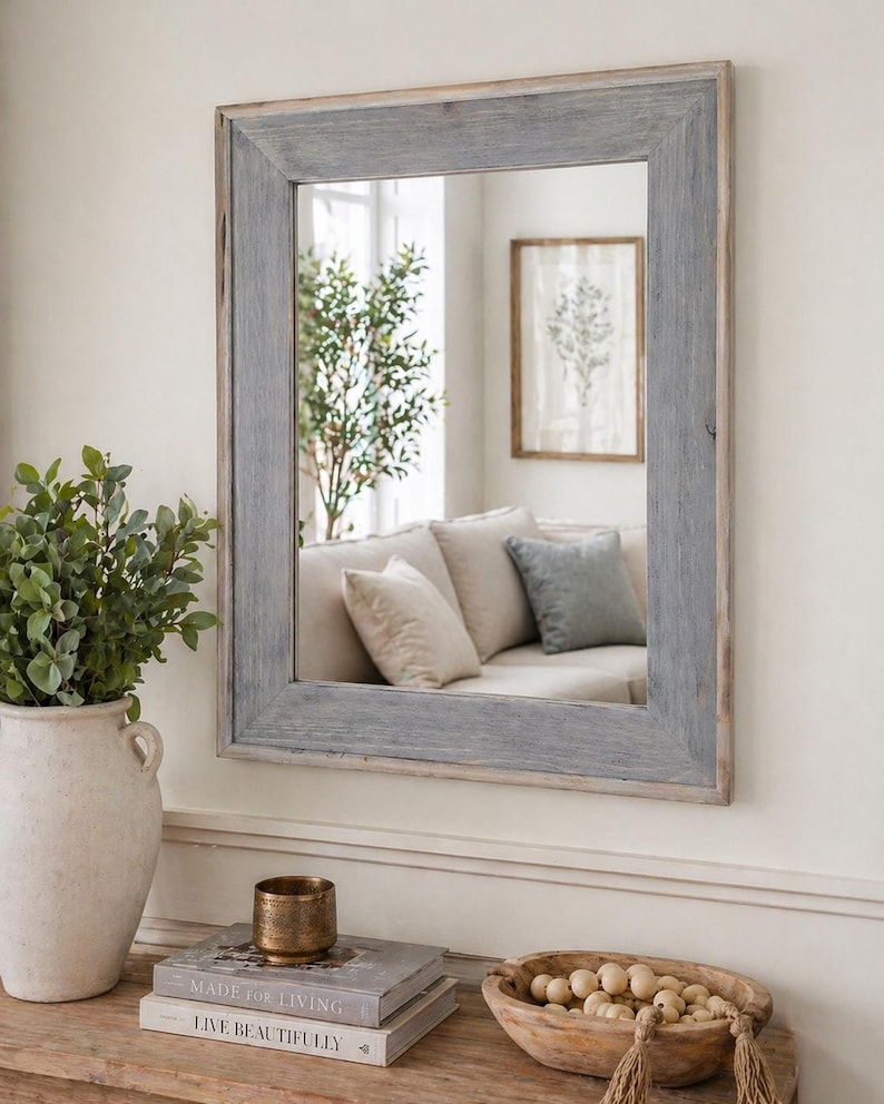 May include: A rectangular mirror with a grey wooden frame hangs on a white wall. The mirror reflects a living room with a sofa and artwork. A vase with greenery, books, and a decorative bowl sit on a wooden table below the mirror.