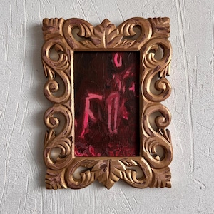 May include: A rectangular picture frame with an ornate, carved gold finish featuring scrollwork and leaf motifs. The frame contains a dark, textured painting with abstract, bright pink brushstrokes and markings that resemble the letters F and O.