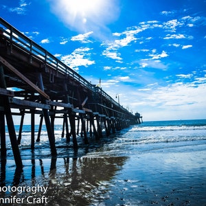 May include: A long wooden pier extends into the ocean under a bright blue sky with scattered clouds. The pier's structure is reflected in the wet sand, creating a symmetrical image. The sun shines brightly, adding to the coastal scene.