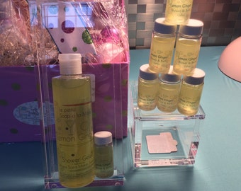 May include: A set of six clear glass bottles filled with a yellow liquid. The bottles are labeled "Lemon Ginger Shower & Bath Gel" and are arranged in a pyramid shape on a clear acrylic shelf. The bottles are in front of a pink and white gift box.