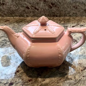 May include: A pink ceramic teapot with a square lid and a decorative handle. The teapot has a spout and a lid with a small knob. The teapot is sitting on a granite countertop.