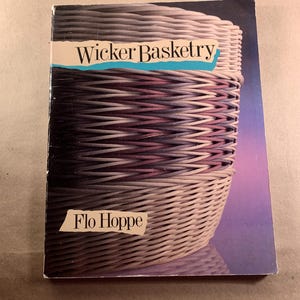 May include: A book titled "Wicker Basketry" by Flo Hoppe. The cover features a close-up image of a woven basket in shades of purple, gray, and white. The title is in a bold, sans-serif font with a blue background.
