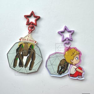 May include: Two acrylic keychains with star-shaped clasps. The left keychain features a brown creature inside a geometric sphere with the text SAVIOR OF THE UNIVERSE. The right keychain shows a blonde character sitting next to a similar sphere.