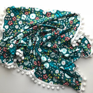 May include: A teal blue fabric baby blanket with a white pom-pom trim and a floral pattern in red, yellow, white, and green.