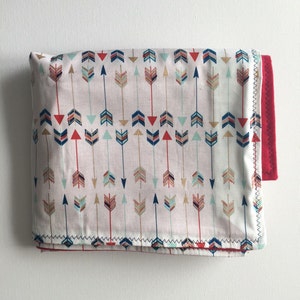 May include: A folded baby blanket with a white background and a pattern of colorful arrows. The blanket has a red backing.