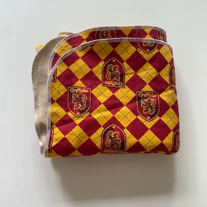 May include: A red and yellow fabric blanket with a Gryffindor house crest pattern. The blanket is folded in half and has a white fleece lining.