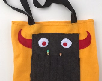 May include: A yellow felt tote bag with a black felt monster face. The monster has red horns, white eyes, and a black felt pocket with two crayons sticking out.