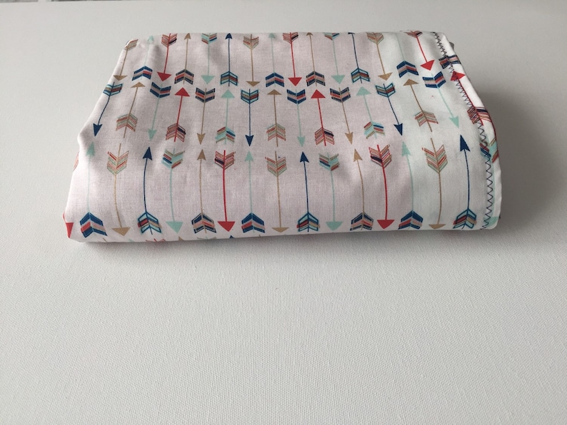 May include: A folded fabric with a white background and a repeating pattern of colorful arrows. The arrows are in shades of red, blue, green, and gold.