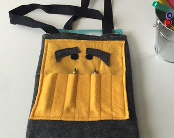 May include: A yellow felt crayon holder with a black strap. The holder has six pockets for crayons and a felt face with black eyebrows and black eyes.