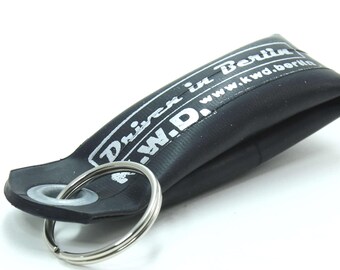 May include: A black rubber keychain loop featuring a silver metal ring and a white printed graphic that reads Driven in Berlin and W.D. with the website address www.kwd.berlin. The material appears to be recycled bicycle inner tube rubber.