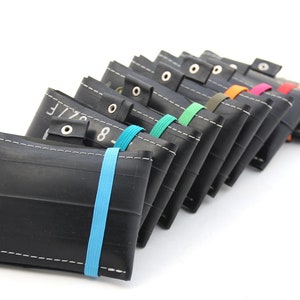 May include: A row of black recycled tire wallets with colorful elastic bands. Each wallet has a white stitch detail and a metal snap closure.