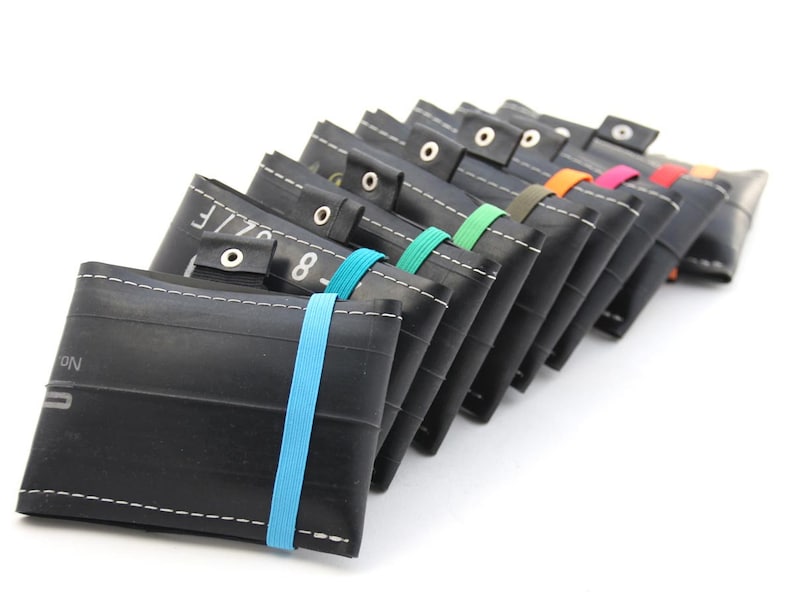May include: A row of black recycled tire wallets with colorful elastic bands. Each wallet has a white stitch detail and a metal snap closure.