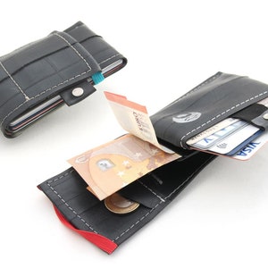 May include: Two black recycled tire wallets, one open to show cash and cards, the other closed. The open wallet has a red elastic band and a white stitched seam. The closed wallet has a blue elastic band and a silver snap.
