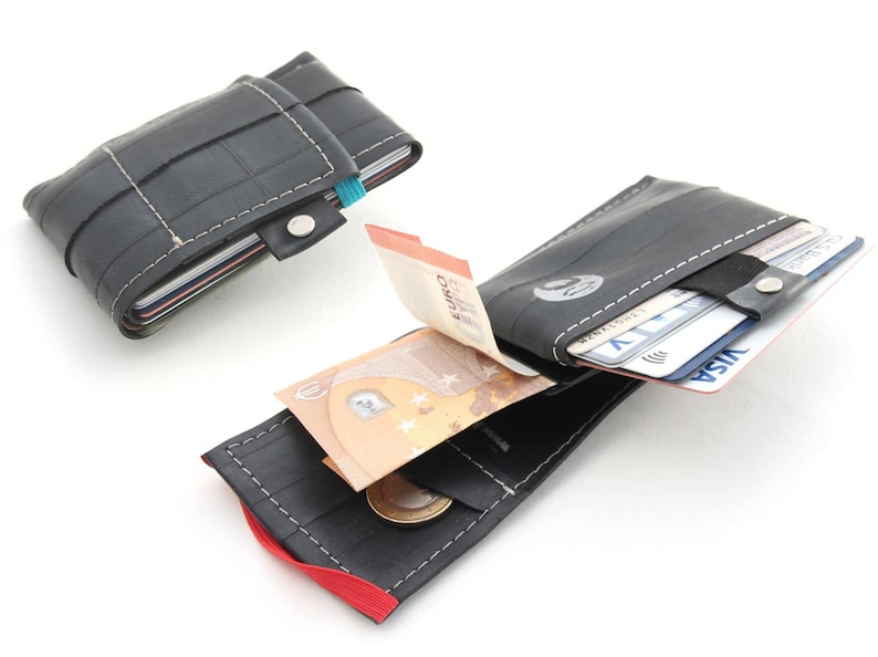 May include: Two black recycled tire wallets, one open to show cash and cards, the other closed. The open wallet has a red elastic band and a white stitched seam. The closed wallet has a blue elastic band and a silver snap.