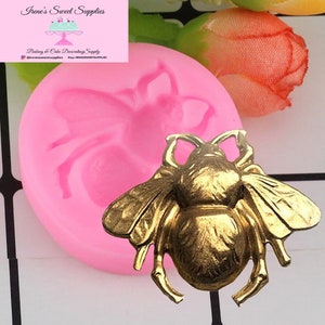 Peut inclure: Un moule en silicone rose en forme d'abeille et un ornement d'abeille doré. Le moule est rond et présente un motif d'abeille détaillé. L'abeille dorée a des ailes et des détails du corps complexes. L'image comprend également le texte "Irene's Sweet Supplies".